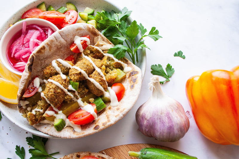 A brightly lit picture of a falafel wrap with toasted Arabic flatbread, hummus, cucumbers, tomato, and falafel with tahini drizzle on top next to a dish of pickled onions, parsley and cucumber salad.