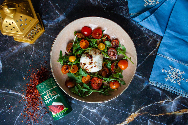 A picture of a bowl of burrata salad with arugula, cherry tomatoes and an Aleppo Pepper vinaigrette on a dark counter next to a container of Afia Aleppo Pepper Mediterranean pantry Spice