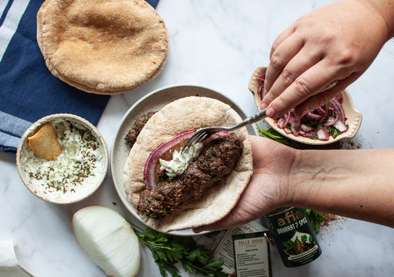 A picture of a hand holding a pita wrap made with kofta kebabs, sumac onions and yogurt dip on a white marble counter next to a container of Afia Baharat 7 Spice Mediterranean Pantry Spice