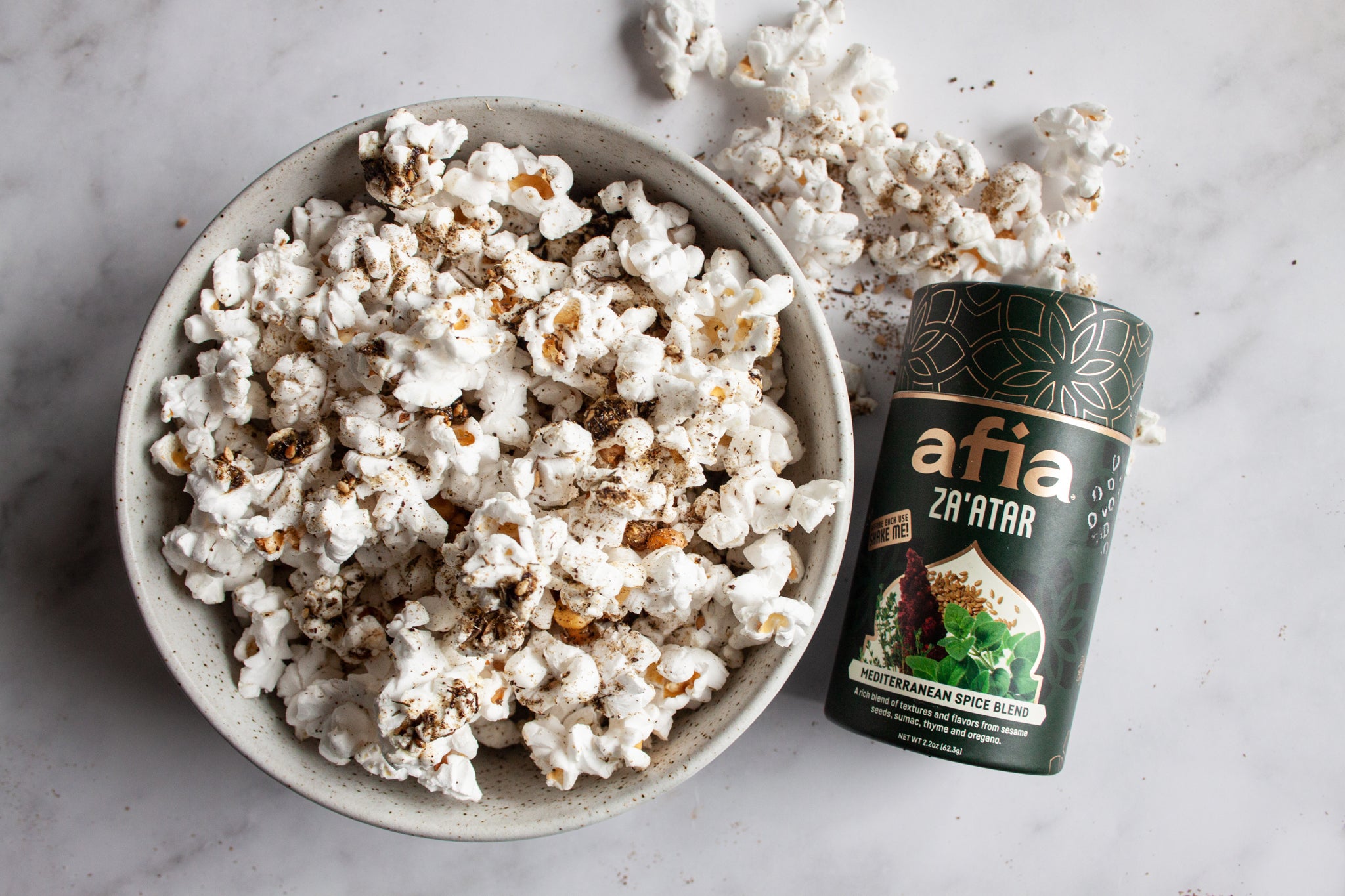 A picture of a bowl of popcorn topped with za'atar spice mix next to a container of Afia Za'atar Mediterranean Spice Blend and a gold lantern